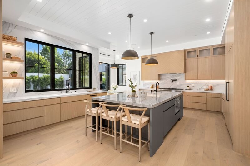 Interior de cocina moderna con una gran isla, gabinetes de madera y lámparas colgantes, que cuenta con una gran ventana con vista a la vegetación.
